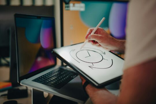 person in front of a laptop drawing a clock on a tablet