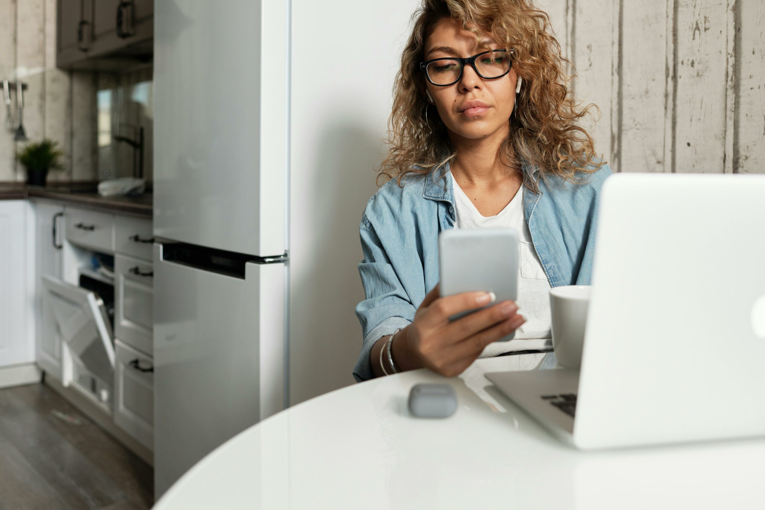 Woman working at a kitchen table with a cellphone and a laptop. Behind her we can see kitchen appliances like a fridge and a dishwasher, probably all connected with IoT